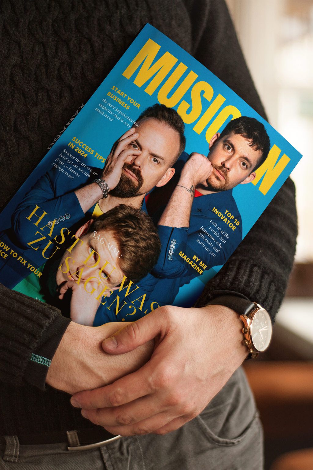 Eine Person, die einen schwarzen Pullover und eine Uhr trägt, hält ein Musician-Magazin in der Hand, auf dessen Titelseite zwei Männer abgebildet sind, von denen einer mit der Hand auf seinem Gesicht posiert. Die Zeitschrift hat ein leuchtend blaues und gelbes Design.