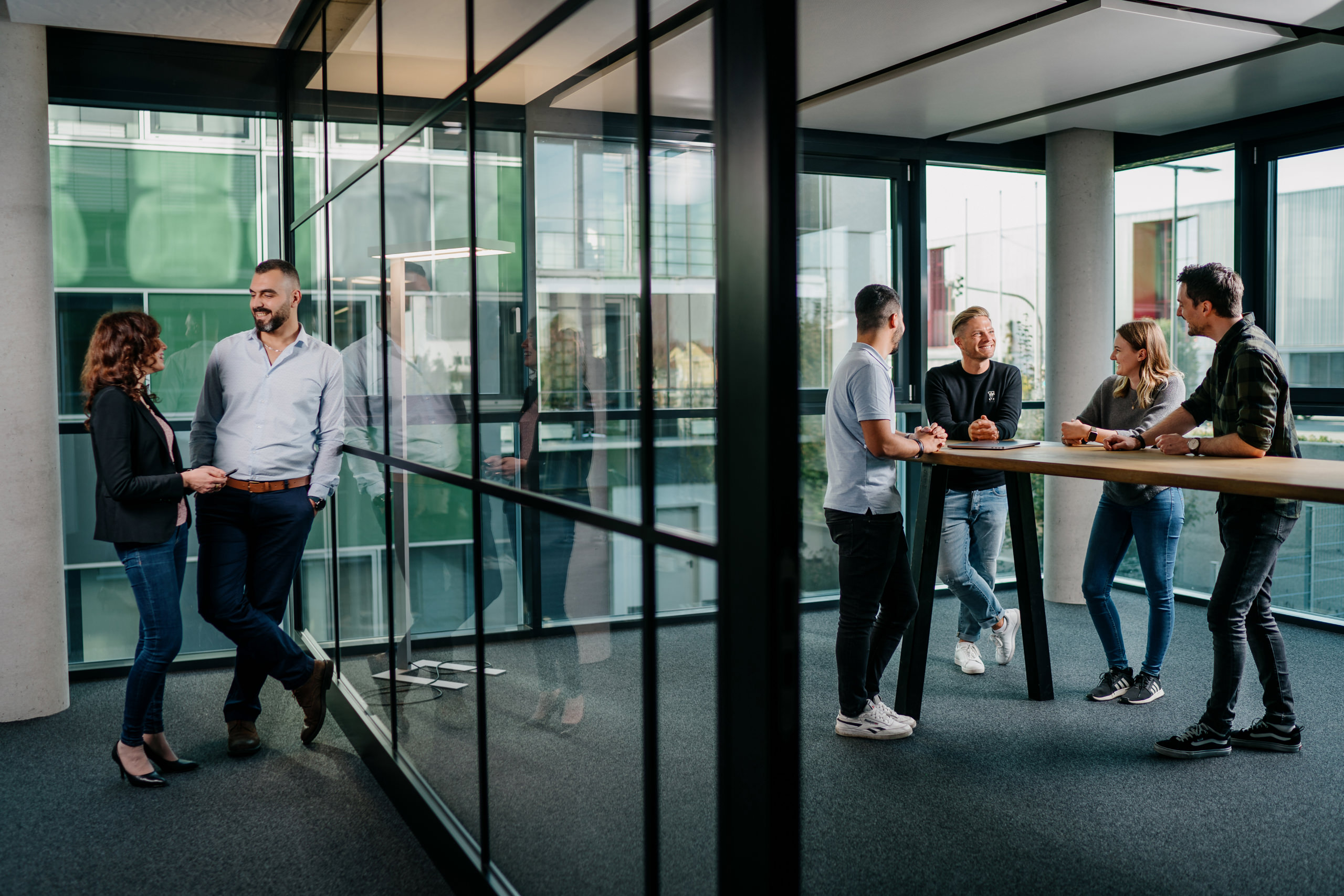 Zwei Gruppen von Personen in legerer Geschäftskleidung unterhalten sich in einem modernen Büro mit großen Fenstern und Glaswänden; ein Paar steht in der Nähe des Fensters, und vier andere unterhalten sich an einem hohen Tisch.
