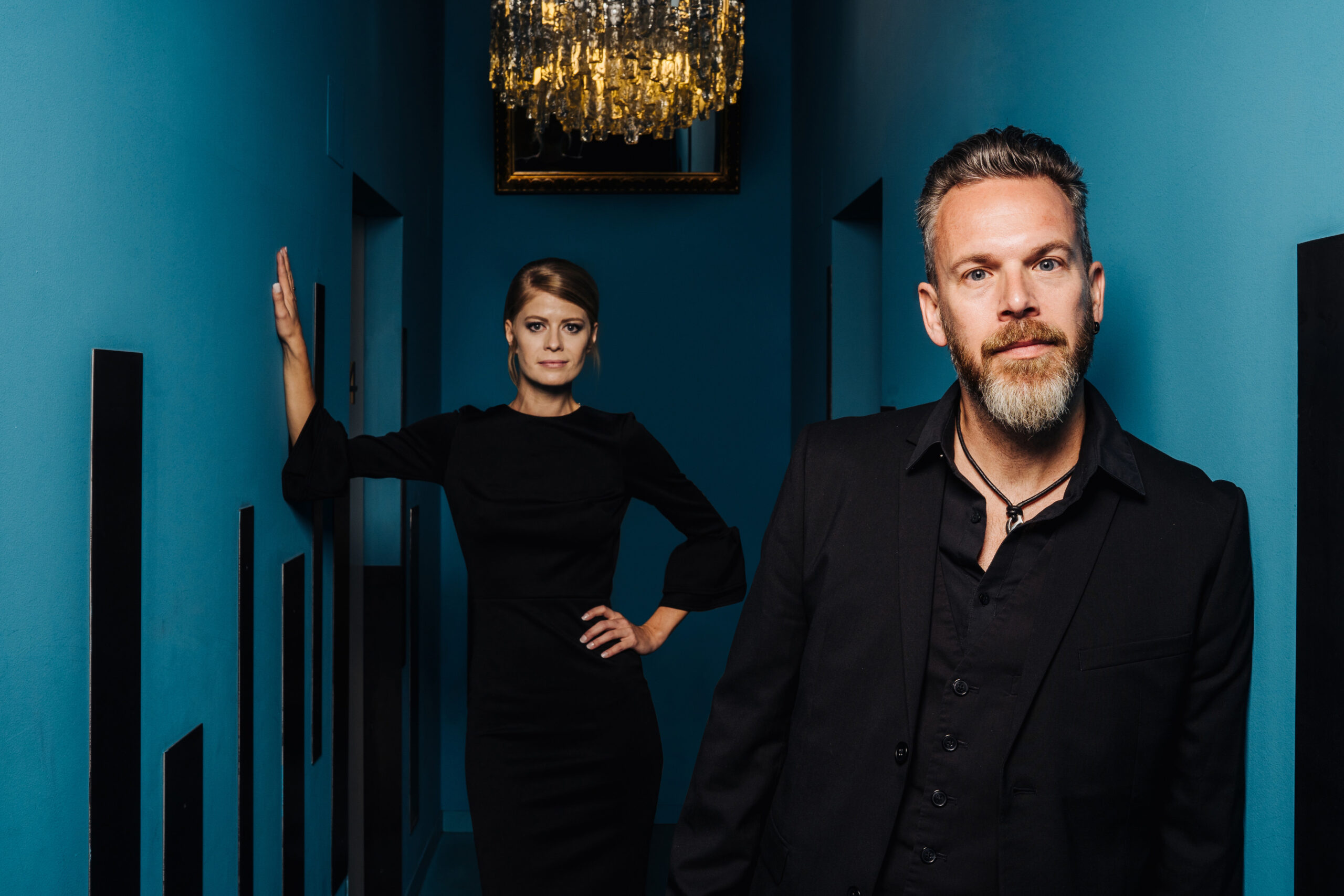 Im Vordergrund steht ein bärtiger Mann in einem schwarzen Anzug, im Hintergrund eine Frau in einem schwarzen Kleid, die eine Hand in die Hüfte gestützt hat. Beide stehen in einem schmalen Flur mit blauen Wänden und einem Kronleuchter darüber.