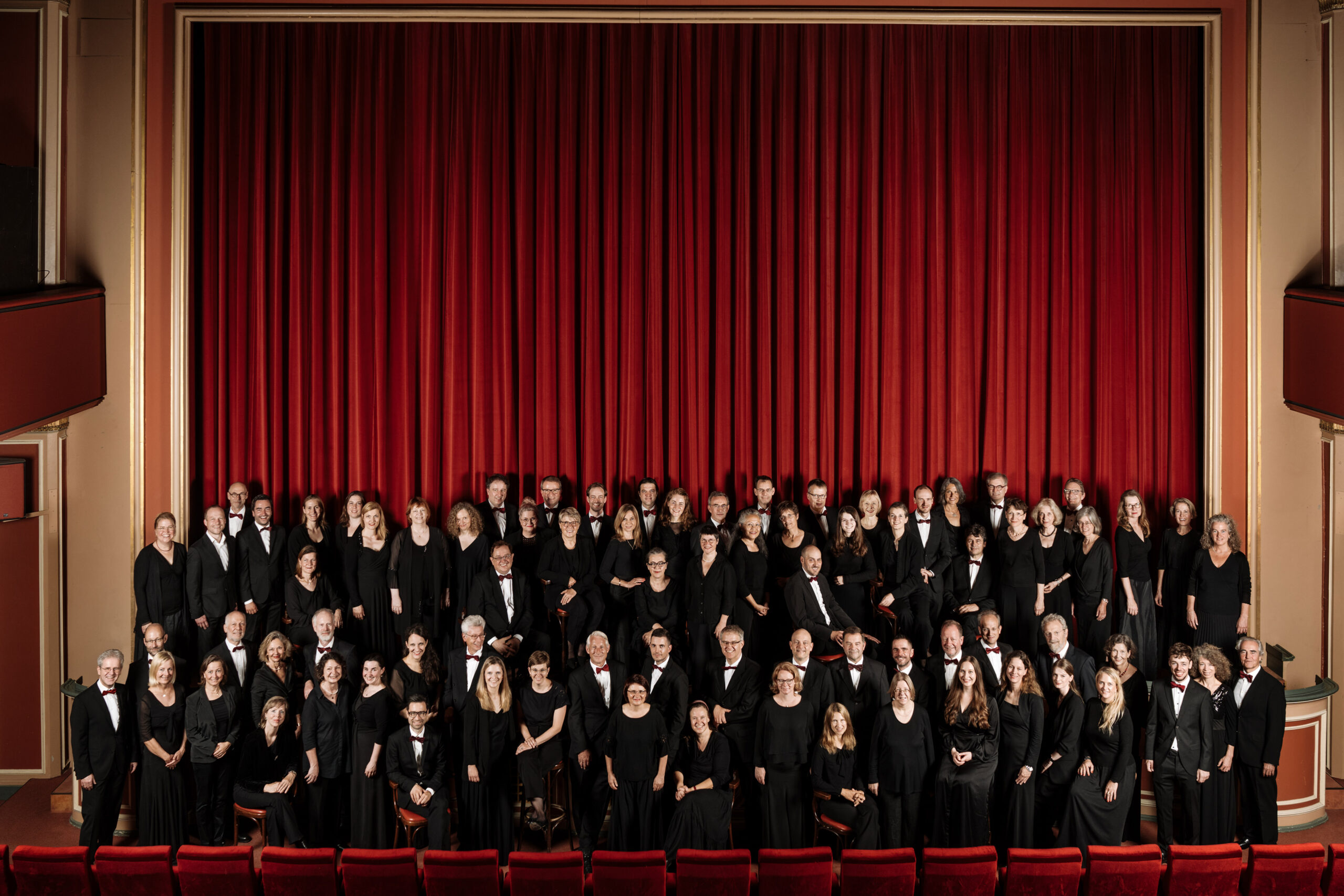 Ein großes Orchester in formeller schwarzer Kleidung steht auf der Bühne vor einem roten Vorhang und posiert für ein Gruppenfoto in einem Theater mit roten Wänden und Sitzen.