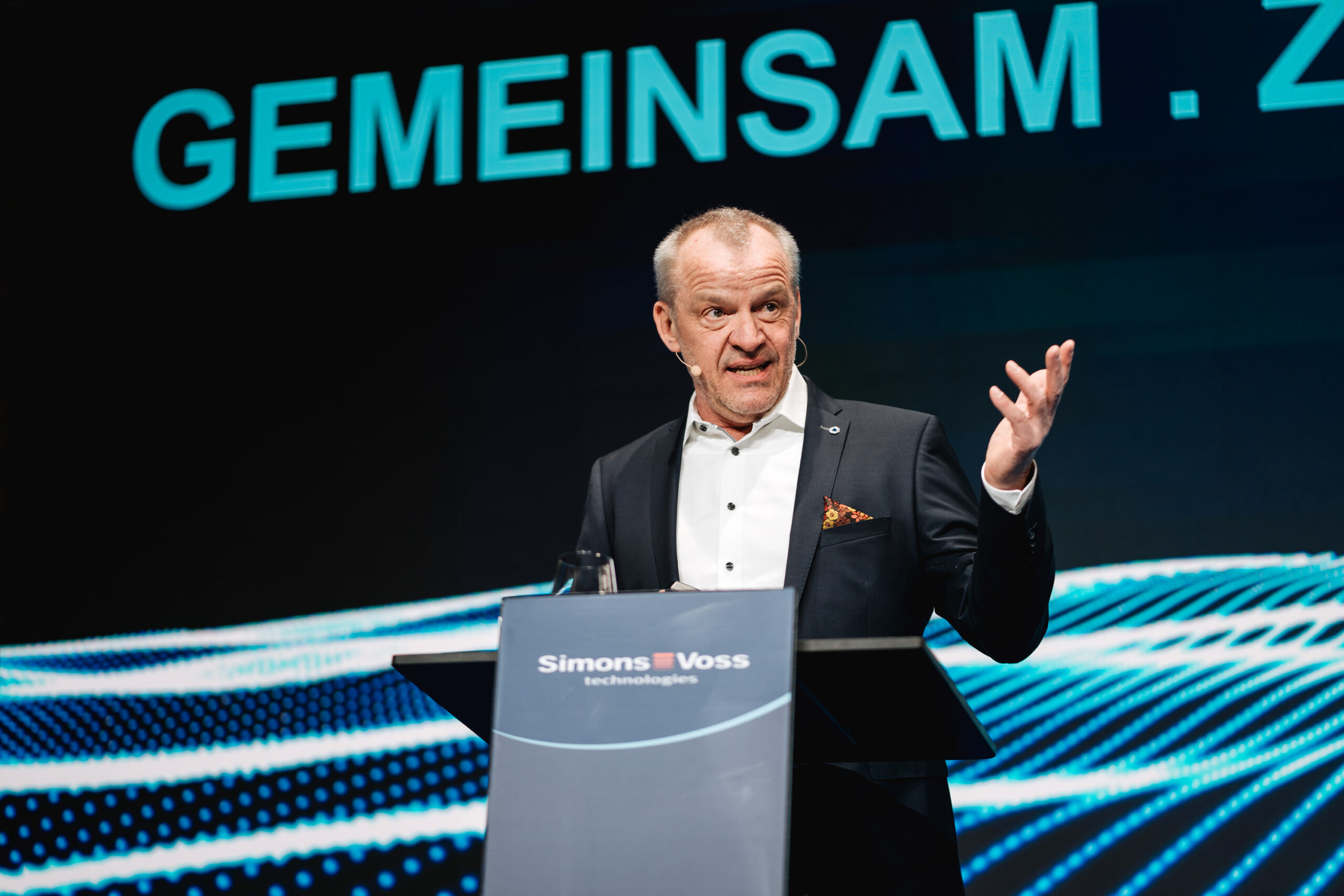 Ein Mann im Anzug spricht an einem Podium mit der Aufschrift SimonsVoss technologies und gestikuliert mit einer Hand. Hinter ihm erscheint das Wort GEMEINSAM auf einem großen Bildschirm mit einem blauen digitalen Muster.