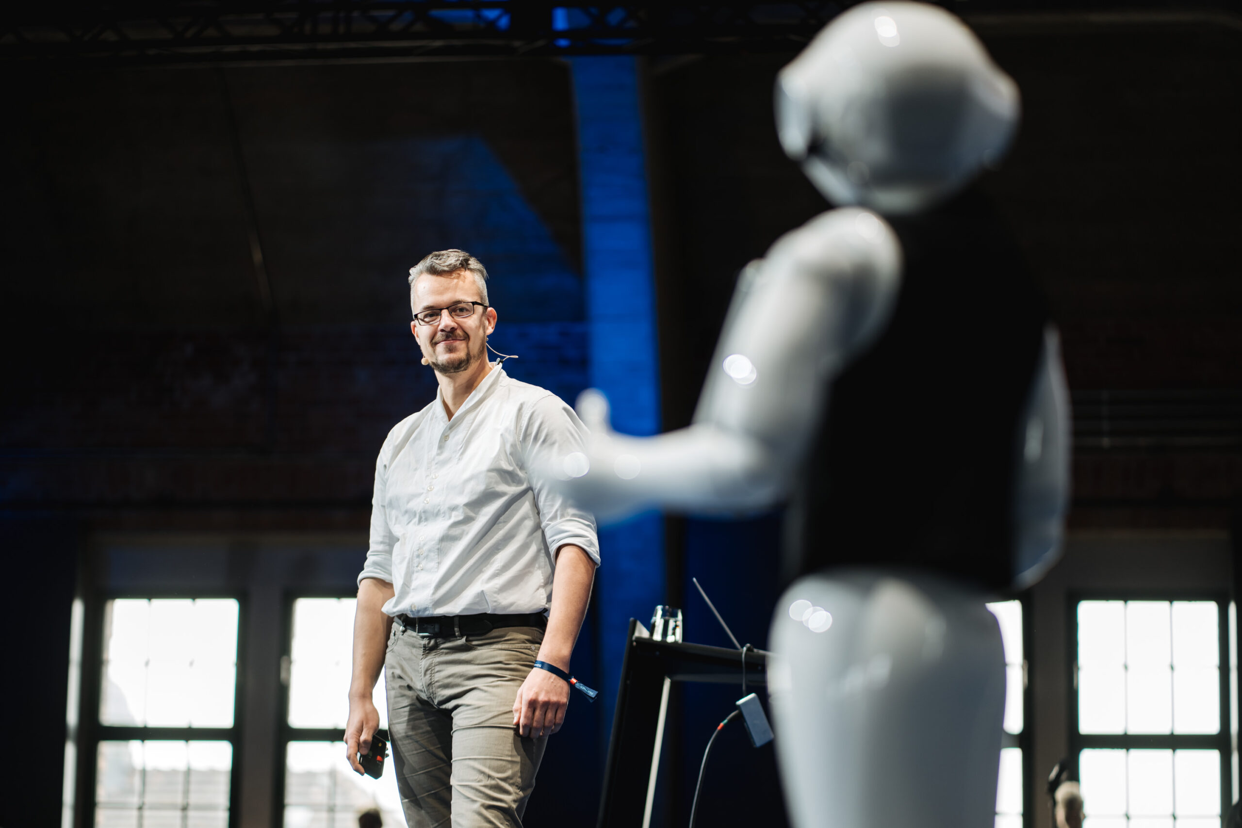Ein Mann, der eine Brille und ein Headset trägt, steht auf der Bühne und blickt auf einen humanoiden Roboter, der ihm gegenübersteht. Der Schauplatz scheint eine moderne, industriell geprägte Veranstaltung oder Konferenz mit großen Fenstern im Hintergrund zu sein.
