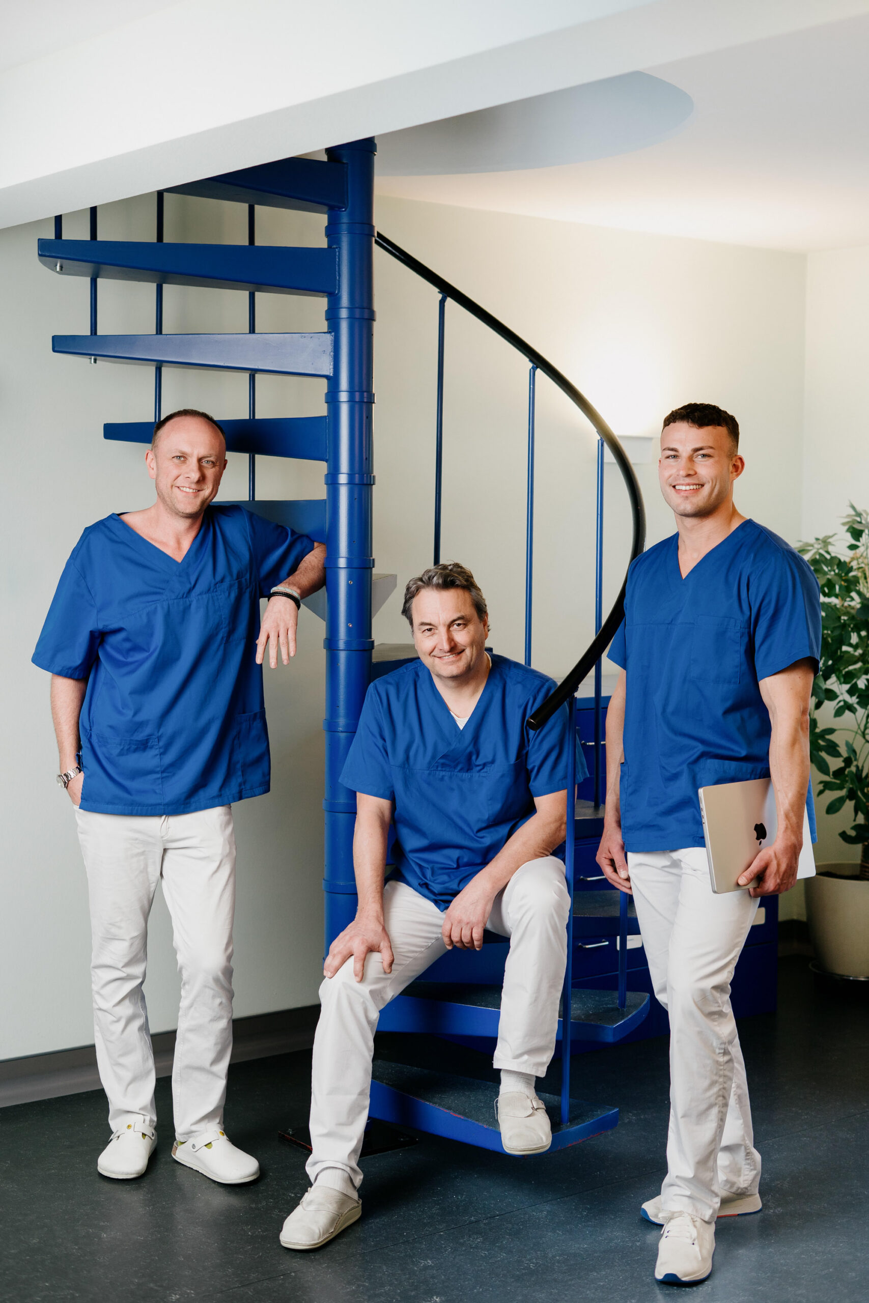 Drei Männer in blauen Arztkitteln und weißen Hosen posieren vor einer blauen Wendeltreppe. Einer sitzt auf einer Stufe und hält ein Tablet in der Hand, die beiden anderen stehen lächelnd in einem hellen, modernen Raum.