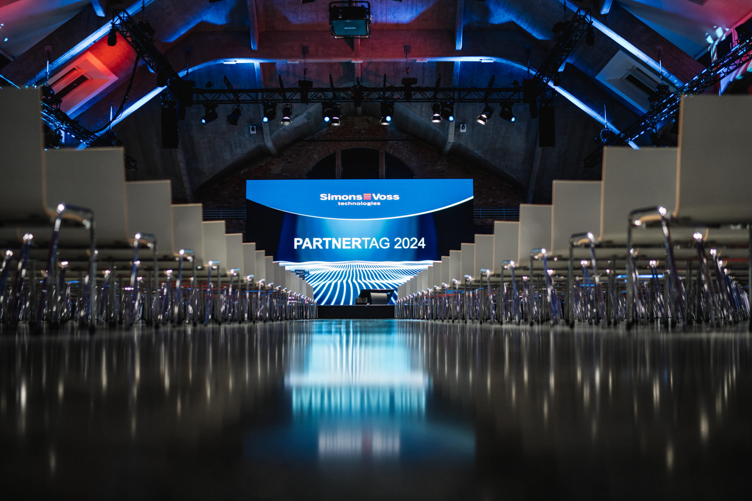 Ein großer Veranstaltungssaal mit leeren Stuhlreihen vor einer hell erleuchteten Bühne, auf der der SimonsVoss PARTNERTAG 2024 auf einer Leinwand unter dramatischer blauer und weißer Beleuchtung zu sehen ist.