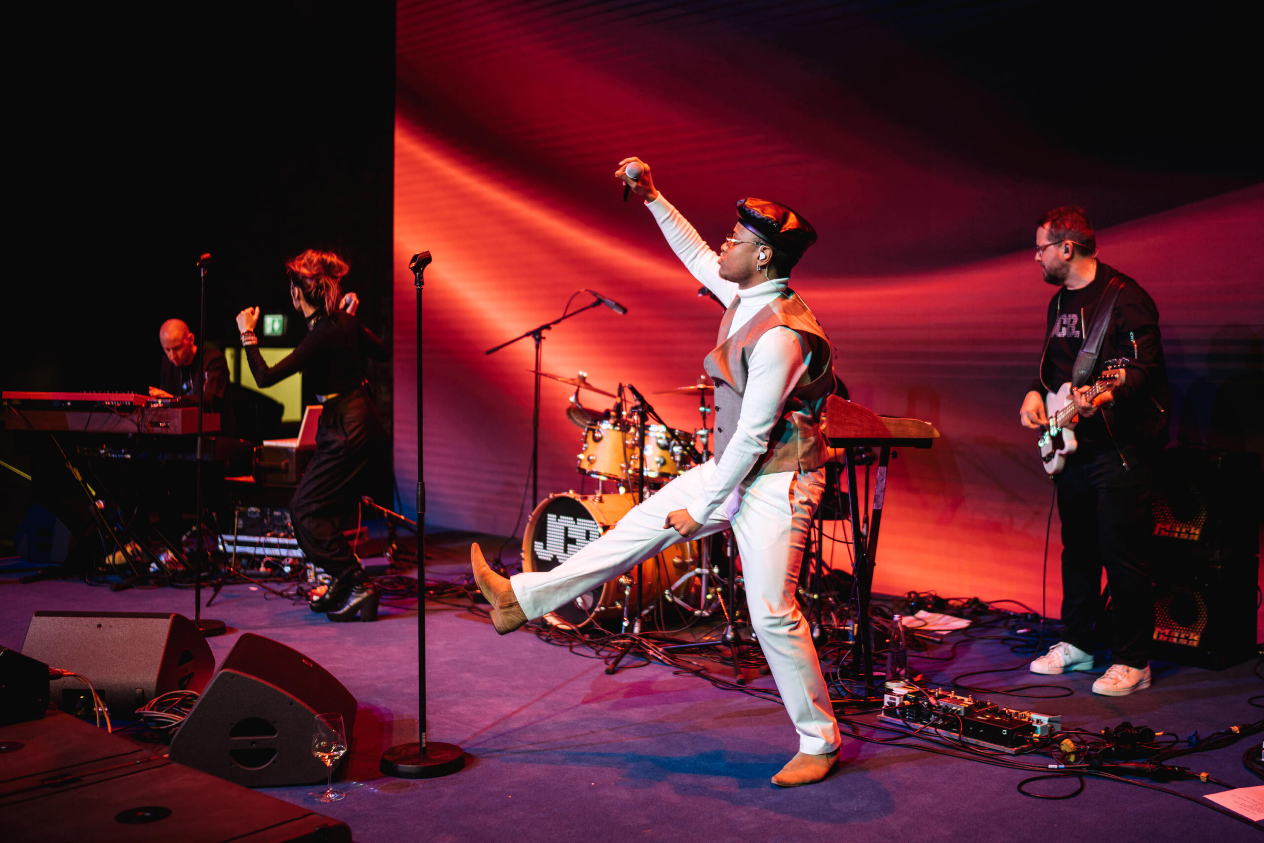Eine lebhafte Band spielt auf der Bühne unter leuchtenden roten und lila Lichtern. Der Leadsänger in weißem Outfit und Hut tanzt energisch in der Mitte, während im Hintergrund Musiker Keyboard, Schlagzeug und Gitarre spielen.