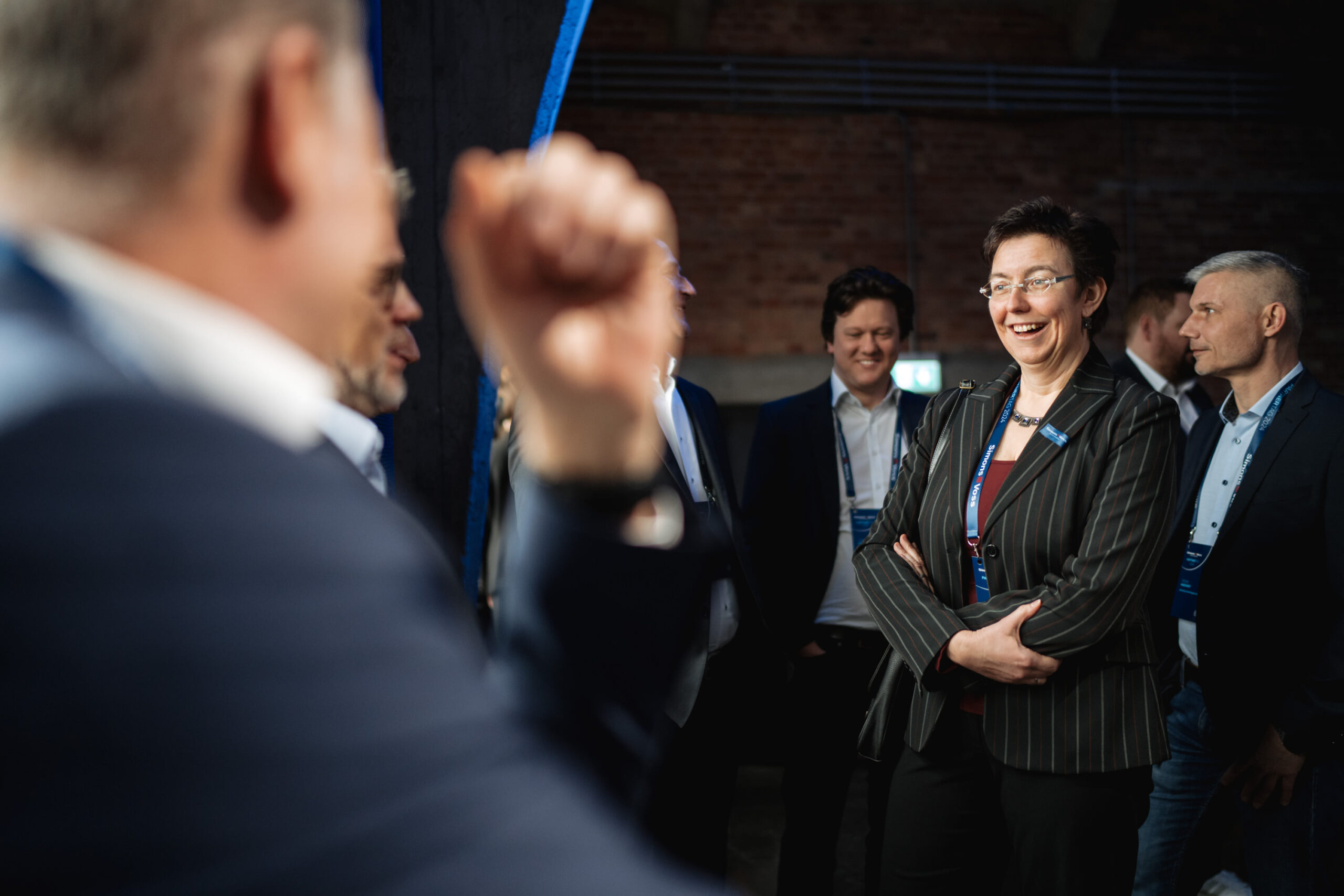 Eine Gruppe professionell gekleideter Personen, darunter eine lächelnde Frau in einem gestreiften Blazer, steht in einem Innenraum zusammen und scheint sich bei einer Geschäftsveranstaltung oder Konferenz angeregt zu unterhalten.