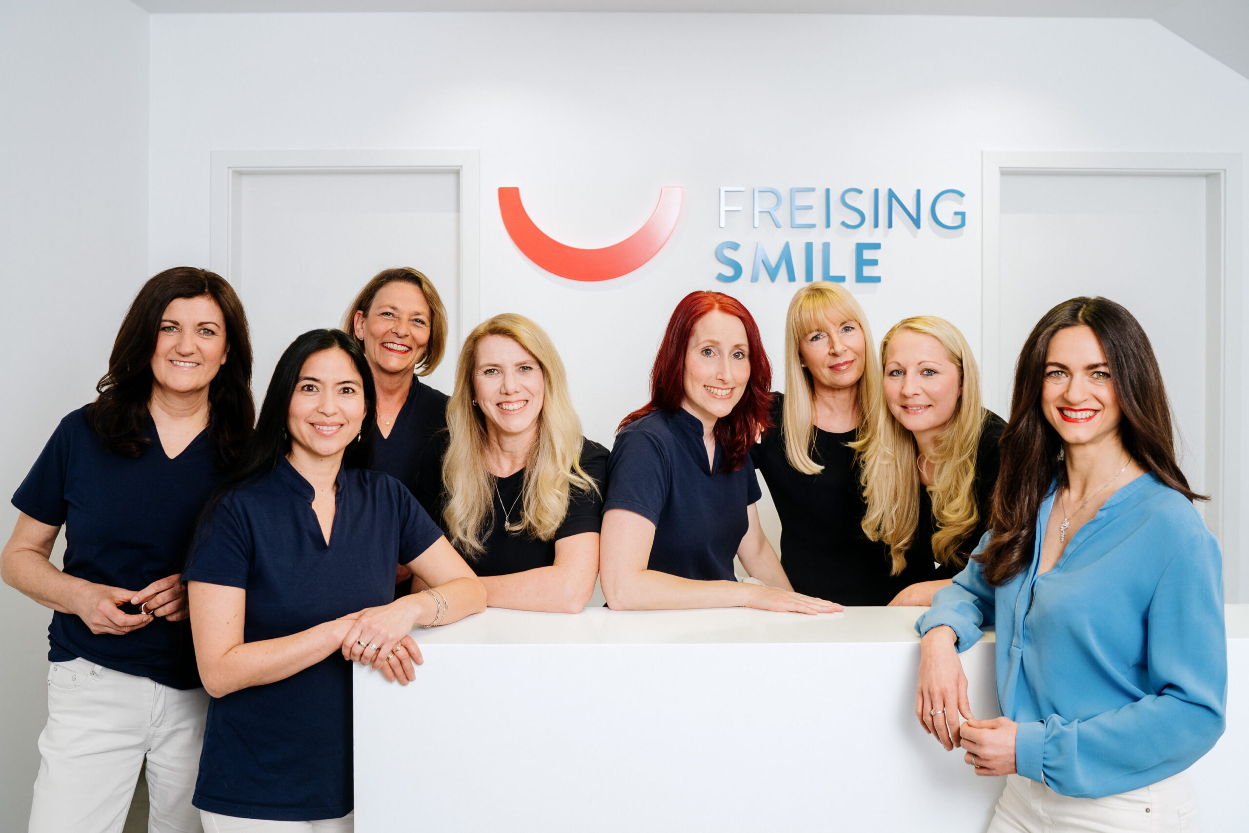Acht Frauen stehen lächelnd hinter einem weißen Empfangstresen mit dem Logo von Freising Smile an der Wand hinter ihnen. Die meisten tragen marineblaue Uniformen, bis auf eine in einer blauen Bluse. Die Umgebung ist sauber und professionell.