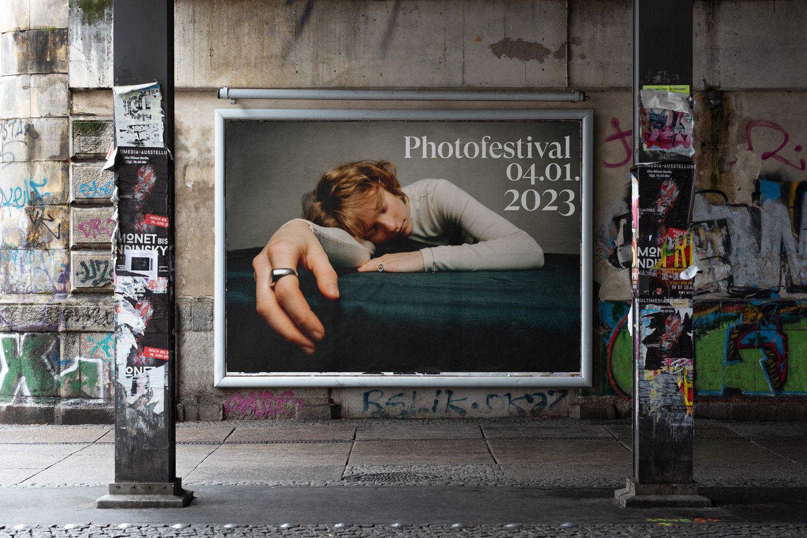 Ein großes Plakat für das Fotofestival 04.01.2023 zeigt eine Person, die ihren Kopf und ihren Arm auf eine dunkle Fläche stützt, angebracht an einer mit Graffiti beschmierten Wand unter einer städtischen Brücke.
