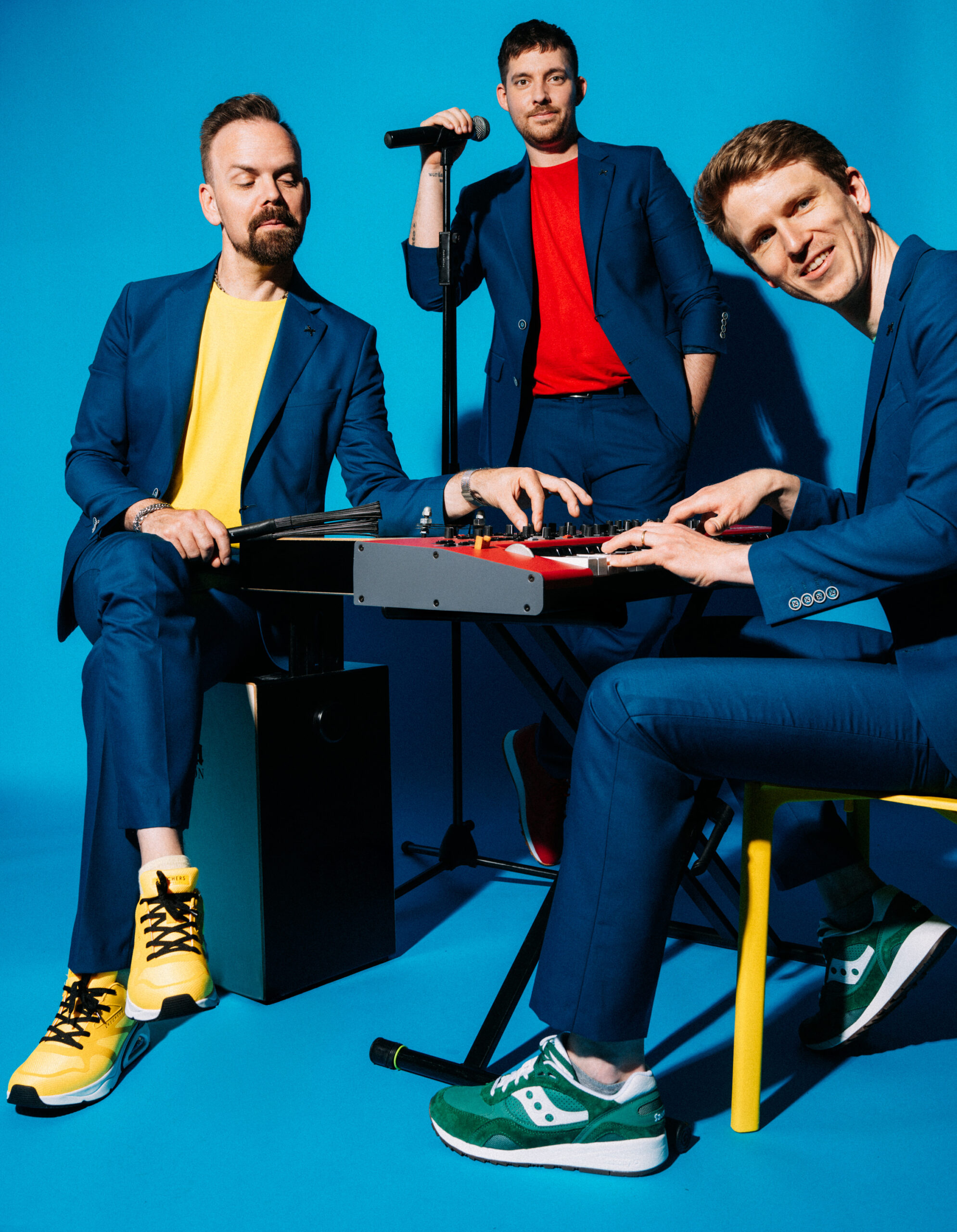 Drei Männer in blauen Anzügen posieren vor einem hellblauen Hintergrund. Einer spielt ein Keyboard, ein anderer sitzt mit Schlagzeugstöcken, und der dritte steht mit einem Mikrofon. Sie tragen bunte Turnschuhe und lässige Hemden in Gelb und Rot.