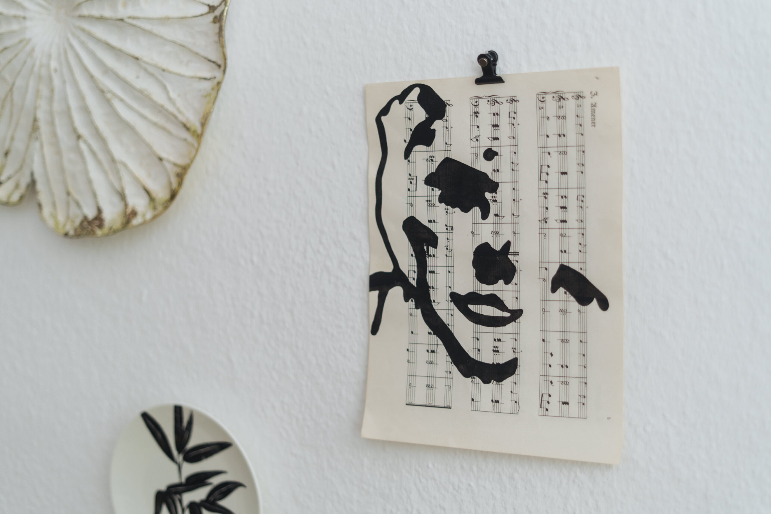 Ein Notenblatt mit einem schwarzen, abstrakten Gesicht ist an eine weiße Wand geklemmt, daneben liegen ein getrocknetes Blatt und ein dekorativer Teller mit einem schwarzen Blattmuster.