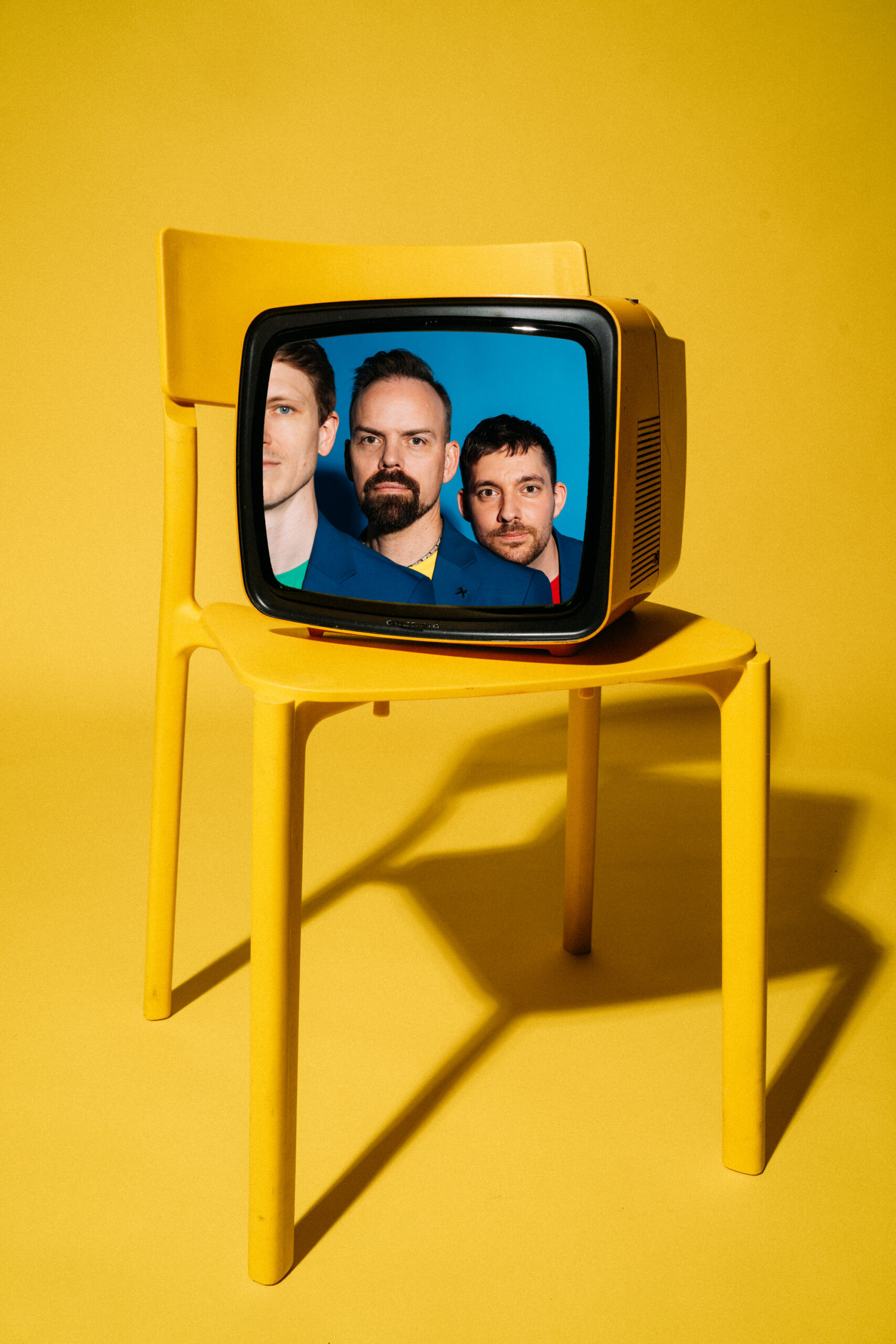 Ein Retro-Fernseher sitzt auf einem gelben Stuhl vor einem gelben Hintergrund und zeigt auf seinem Bildschirm die Gesichter dreier Männer, die jeweils nur teilweise sichtbar sind und hinter denen ein blauer Hintergrund liegt.