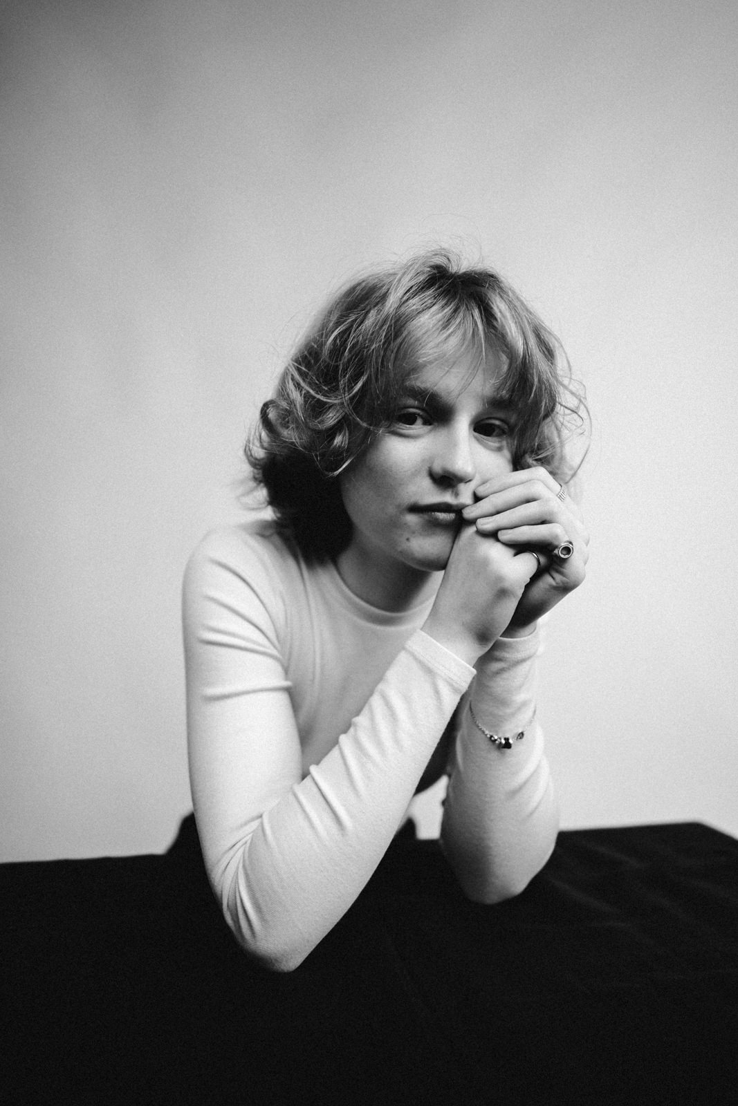 Schwarz-Weiß-Foto einer Person mit gewelltem, schulterlangem Haar, die ein langärmeliges Hemd trägt, die Arme auf einen Tisch stützt und nachdenklich in die Kamera schaut, vor einem schlichten Hintergrund.