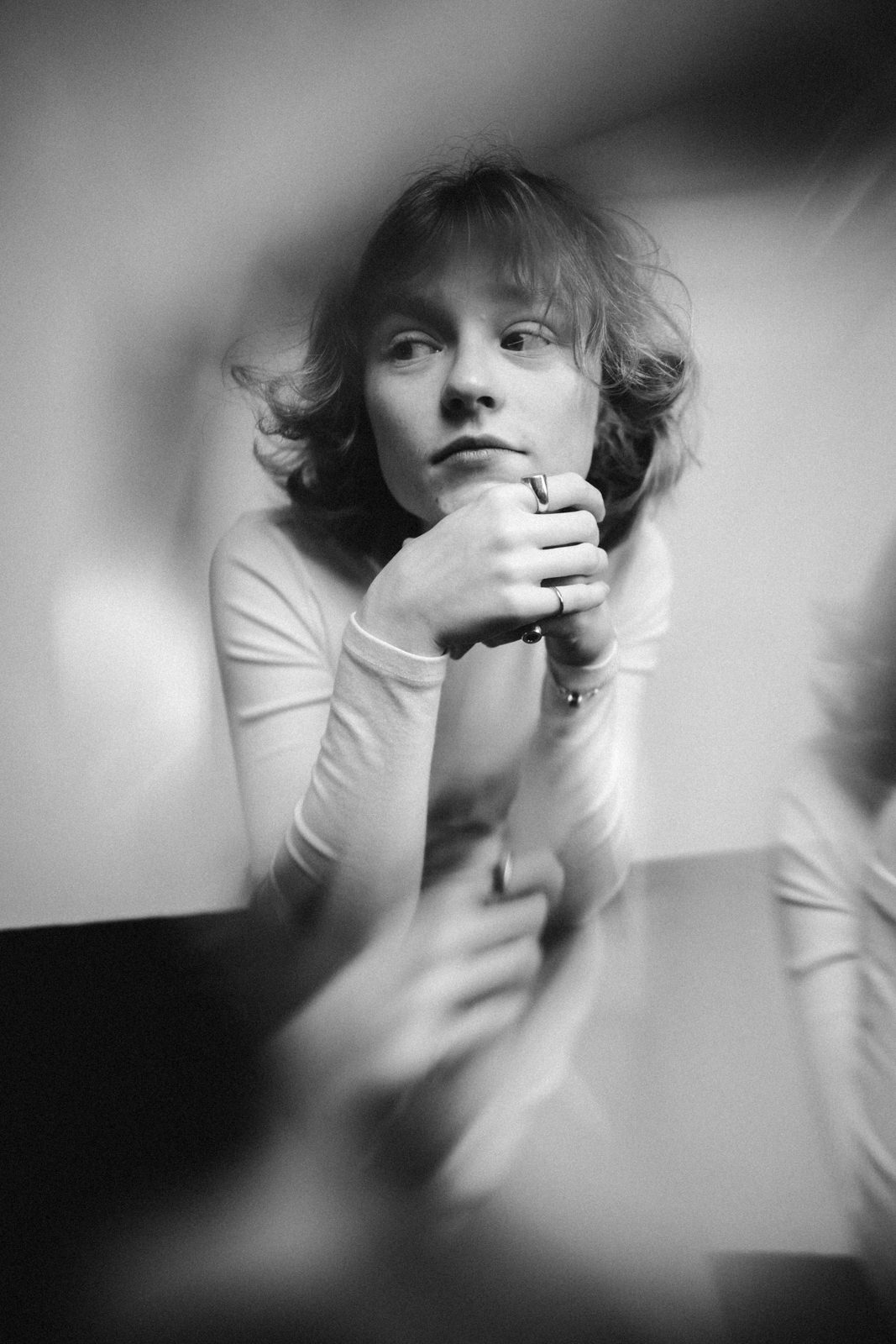 Ein Schwarz-Weiß-Foto einer Person mit kurzem, gewelltem Haar, die nachdenklich zur Seite blickt und ihr Kinn auf die Hände stützt. Das Bild hat einen weichen, verträumten Unschärfeeffekt an den Rändern.