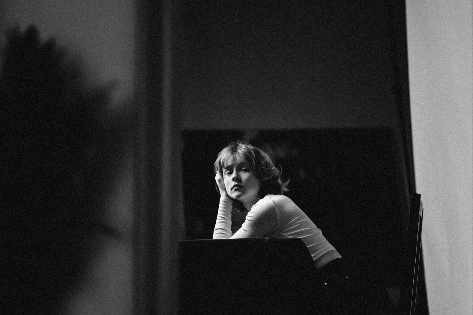 Schwarz-Weiß-Foto einer Person mit kurzen Haaren, die an einem Tisch sitzt, den Kopf auf die Hand stützt und mit nachdenklichem Blick nach vorne blickt. Der Hintergrund ist schummrig und unscharf.