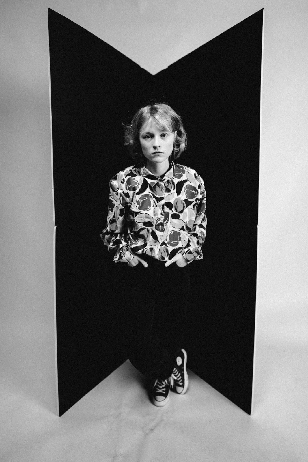 Schwarz-Weiß-Foto einer jungen Person mit kurzen Haaren, die ein gemustertes Hemd, eine dunkle Hose und Turnschuhe trägt und mit den Händen in den Taschen vor zwei großen schwarzen Platten steht, die eine V-Form bilden.