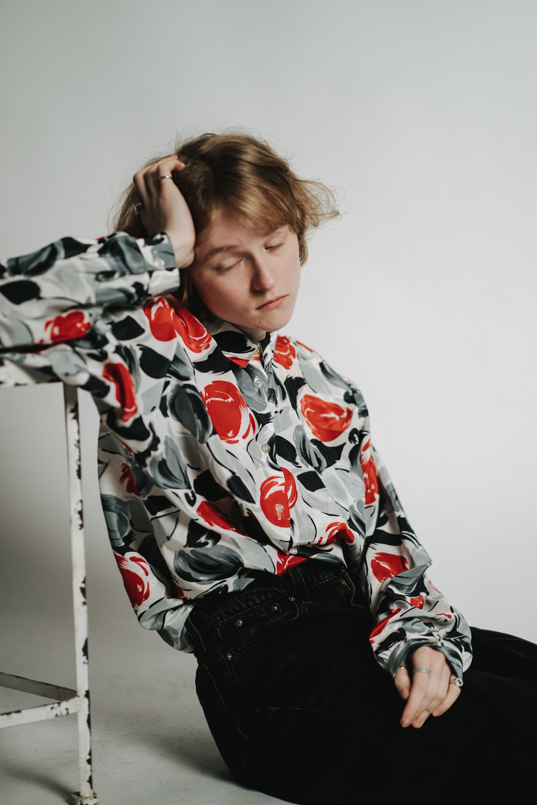 Eine Person mit kurzem, hellbraunem Haar sitzt vor einem weißen Hintergrund, die Augen geschlossen und den Kopf auf eine Hand gestützt. Sie trägt ein gemustertes Hemd mit auffälligen roten, schwarzen und grauen Blumen und eine dunkle Hose.