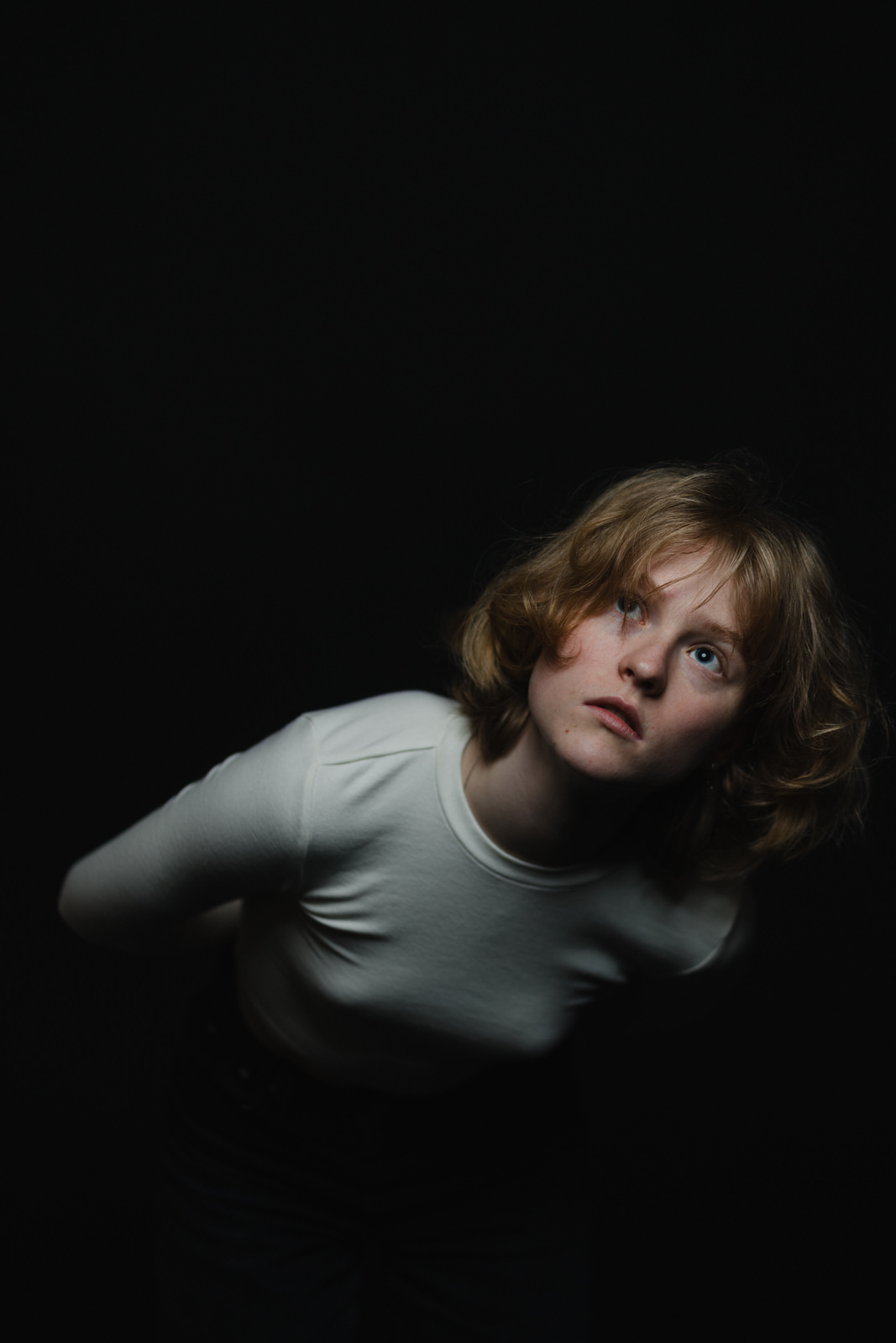 Eine Person mit kurzen, gewellten blonden Haaren, die ein weißes langärmeliges Hemd trägt, beugt sich vor und blickt nach oben vor einem dunklen Hintergrund. Die Beleuchtung hebt ihr Gesicht und ihren Oberkörper hervor.