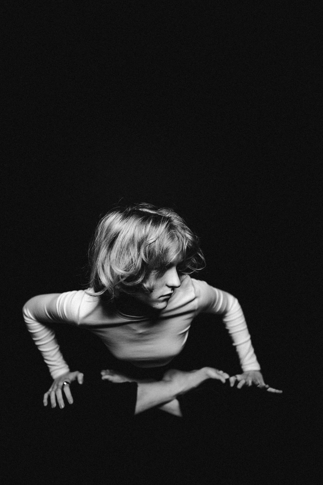 Eine Person mit kurzen Haaren, die ein helles langärmeliges Oberteil trägt, lehnt sich nach vorne, stützt sich mit den Händen auf einer dunklen Fläche ab und blickt nach unten. Das Bild ist in Schwarz-Weiß mit dramatischer Beleuchtung und einem dunklen Hintergrund gehalten.