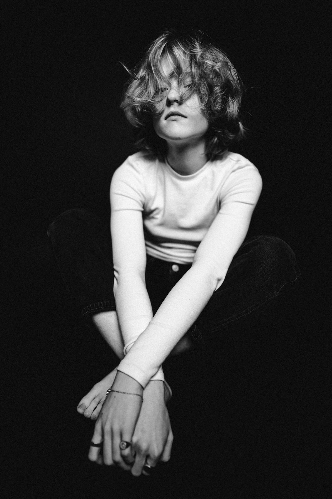 Schwarz-Weiß-Foto einer Person mit gewelltem Haar, die im Schneidersitz sitzt, ein langärmeliges Hemd und Jeans trägt, die Hände vor sich verschränkt und vor einem dunklen Hintergrund leicht nach unten schaut.