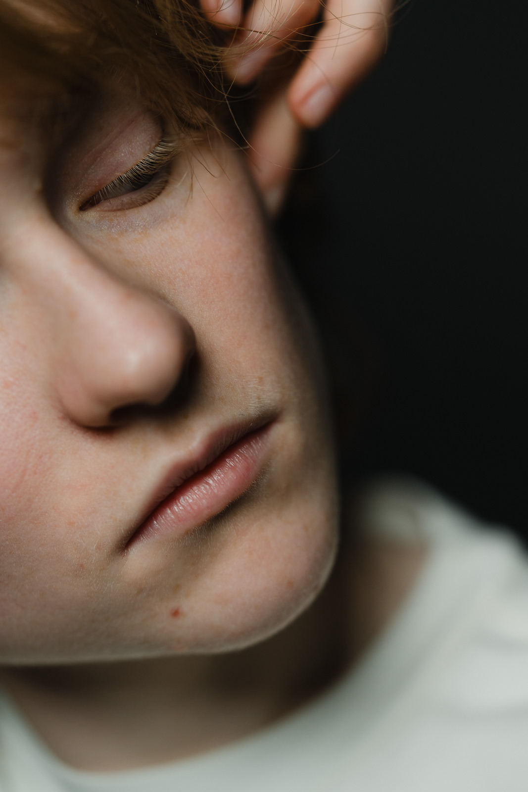 Eine Nahaufnahme des Gesichts einer Person mit geschlossenen Augen, neutralem Ausdruck und heller Haut. Eine Hand berührt sanft die Stirn, die Beleuchtung ist weich und hebt die Struktur der Haut hervor. Der Hintergrund ist dunkel und unscharf.