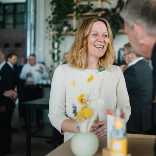 Eine Frau lächelt und unterhält sich mit einem Mann bei einer modernen Indoor-Veranstaltung; im Hintergrund sind andere Menschen und Pflanzen zu sehen, und auf dem Tisch zwischen ihnen steht eine Vase mit gelben und weißen Blumen.