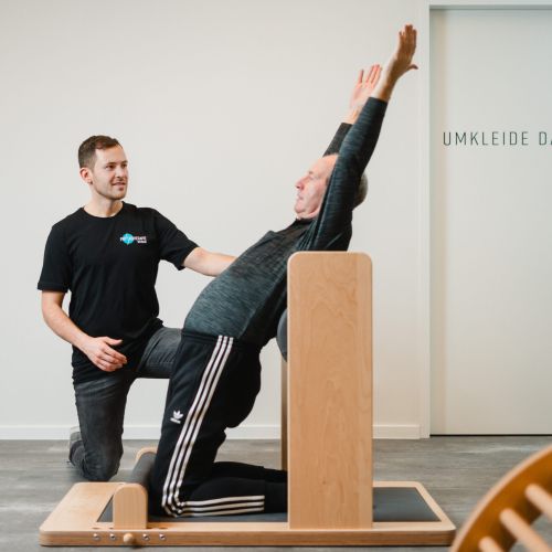 Ein Mann kniet auf einem Trainingsgerät und streckt seine Arme nach oben, während ein Trainer neben ihm kniet und ihn unterstützt. Sie befinden sich in einem Raum in der Nähe einer Tür mit der Aufschrift Umkleide Damen.