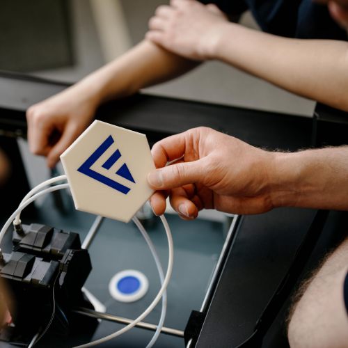 Eine Person hält ein weißes sechseckiges Objekt mit einem blauen geometrischen Logo über einen 3D-Drucker, während andere Personen zusehen; Kabel und Druckerkomponenten sind sichtbar.