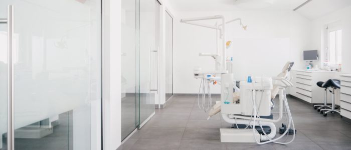 Eine moderne, helle Zahnklinik mit weißen Wänden, Glastrennwänden und zahnmedizinischer Ausrüstung, darunter ein Zahnarztstuhl, Werkzeuge und ein Computer auf einem Tresen im Hintergrund.