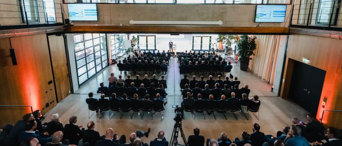 Eine große Gruppe von Personen sitzt in Reihen vor einer Bühne in einem modernen, geräumigen Konferenzsaal und hört einem Redner zu. Der Raum hat hohe Decken, Glaswände und Bildschirme, auf denen Präsentationsfolien zu sehen sind.