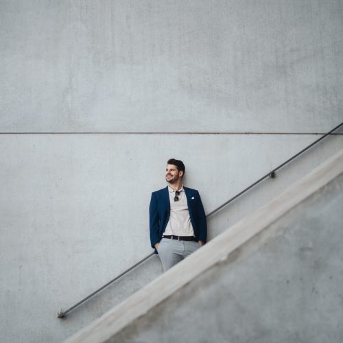 Ein Mann in blauem Blazer und heller Hose steht an einer großen, schlichten Betonwand, die teilweise von einer diagonalen Treppe eingerahmt wird, blickt zur Seite und lächelt, die Hände in den Taschen.