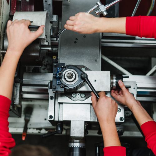 Zwei Personen in roten Ärmeln bedienen eine Metalldrehmaschine von oben und verwenden verschiedene Griffe und Werkzeuge, um ein Metallteil in einer Industrie- oder Werkstatteinrichtung zu bearbeiten.