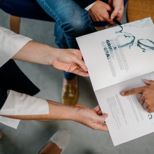 Zwei Personen, die an einem Tisch sitzen, diskutieren über ein gedrucktes Dokument mit Text und Bildern von Brillen, während eine andere Person auf einen Abschnitt des Papiers zeigt. Andere Papiere und ein Brillenetui liegen auf dem Tisch.