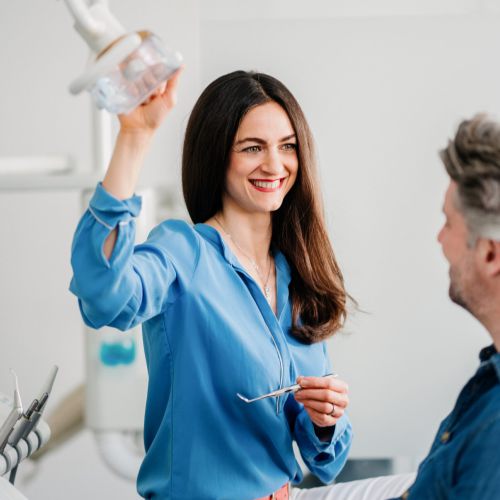 Eine Zahnärztin in einer blauen Bluse lächelt, während sie eine Deckenleuchte für einen männlichen Patienten mit grauem Haar einstellt, der in einer modernen Klinik auf einem Zahnarztstuhl sitzt.
