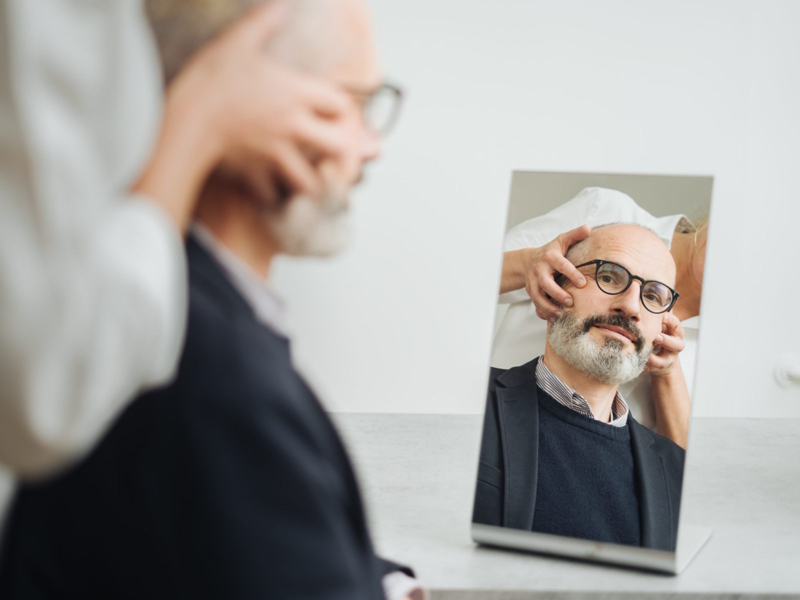 Ein Mann mit Brille und Bart sitzt vor einem Spiegel, während eine andere Person sein Gesicht zurechtrückt, möglicherweise für eine medizinische oder kosmetische Beratung. Das Spiegelbild des Mannes ist im Spiegel deutlich zu erkennen.