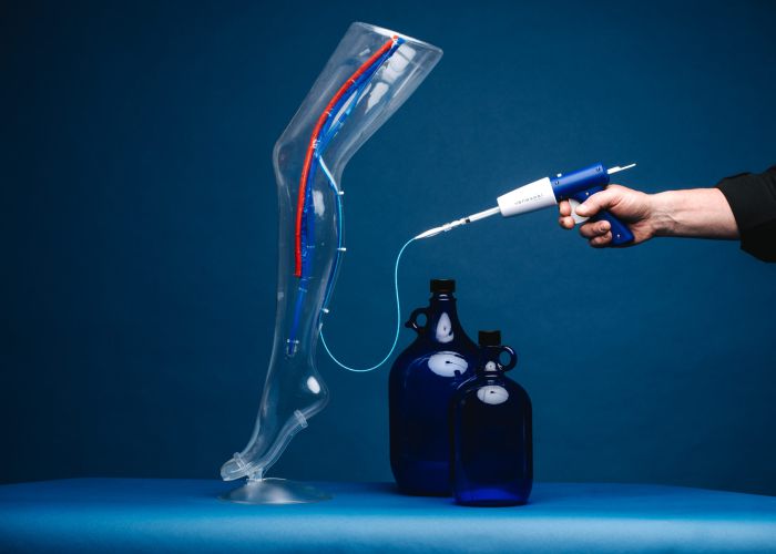Eine Hand hält ein medizinisches Gerät, das durch einen Schlauch mit einem durchsichtigen Kunststoffbeinmodell mit roten und blauen Linien verbunden ist, das neben zwei blauen Glasflaschen auf einer blauen Fläche mit blauem Hintergrund steht.