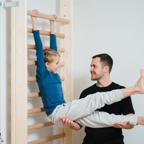 Ein lächelnder Mann stützt einen kleinen Jungen, der an einer hölzernen Sprossenwand hängt, und hält die Beine des Jungen fest, während beide eine spielerische Übung im Haus genießen.