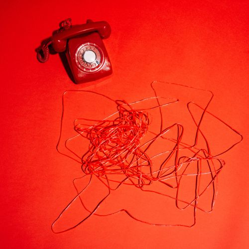 In der linken oberen Ecke einer leuchtend roten Fläche liegt ein rotes Drehtelefon, dessen Kabel sich in einem unordentlichen Haufen in der Nähe verheddert.