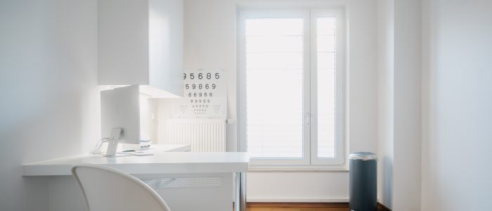 Ein minimalistisches, modernes Augenarztbüro mit weißem Stuhl und Schreibtisch, einer Sehtafel an der Wand, einem Fenster, das natürliches Licht hereinlässt, und einem Mülleimer in der Ecke auf einem Holzboden.