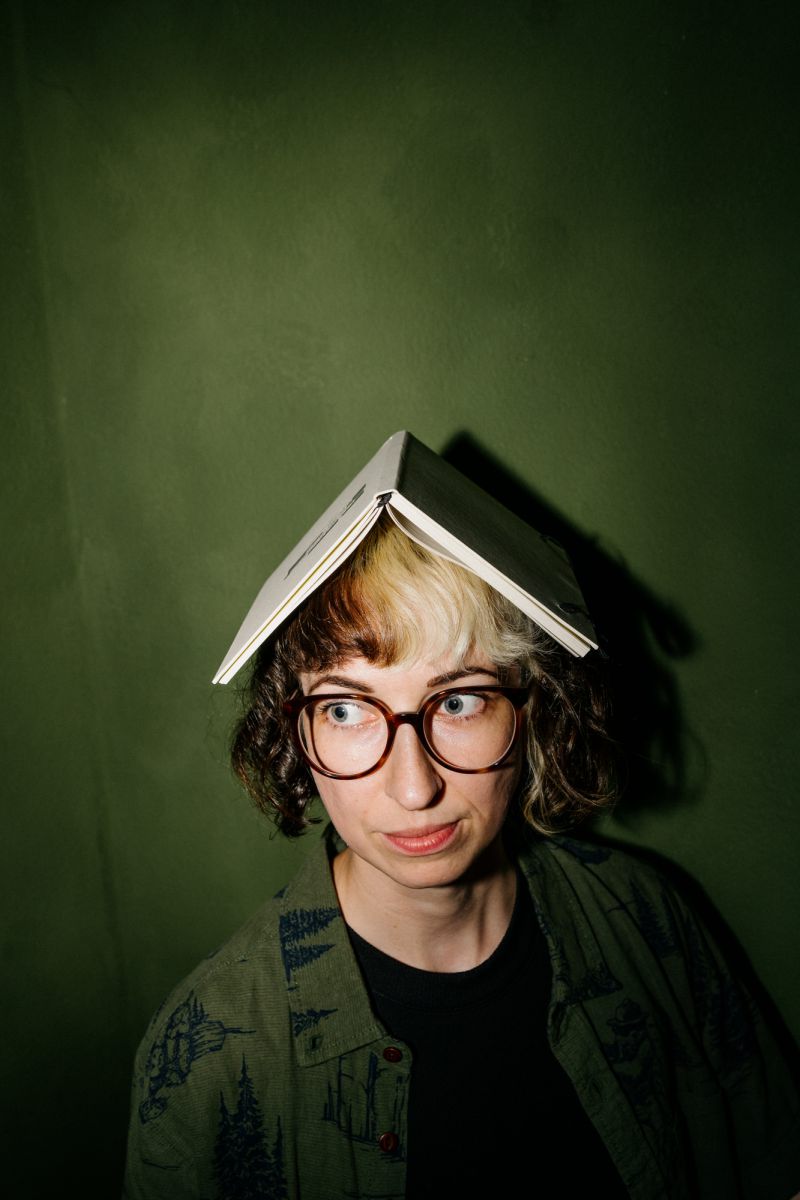 Eine Person mit Brille und kurzem, lockigem Haar hat ein aufgeschlagenes Buch wie einen Hut auf dem Kopf. Sie steht an einer dunkelgrünen Wand und blickt mit nachdenklicher Miene zur Seite.