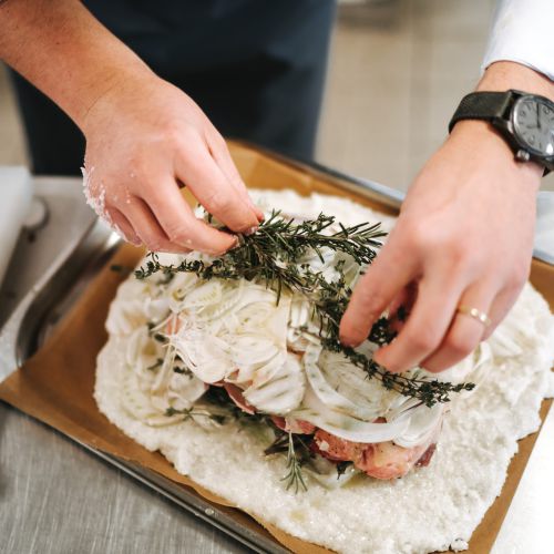 Eine Person legt frische Kräuter auf das mit geschnittenen Zwiebeln bedeckte Fleisch und bereitet es auf einem mit Pergamentpapier und grobem Salz ausgelegten Tablett in einer Küche zu.