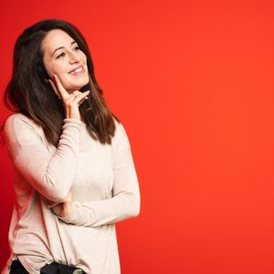Eine Frau mit langen braunen Haaren und einem hellbeigen Pullover steht vor einem leuchtend orangefarbenen Hintergrund, lächelt und stützt ihr Kinn nachdenklich auf ihre Hand.