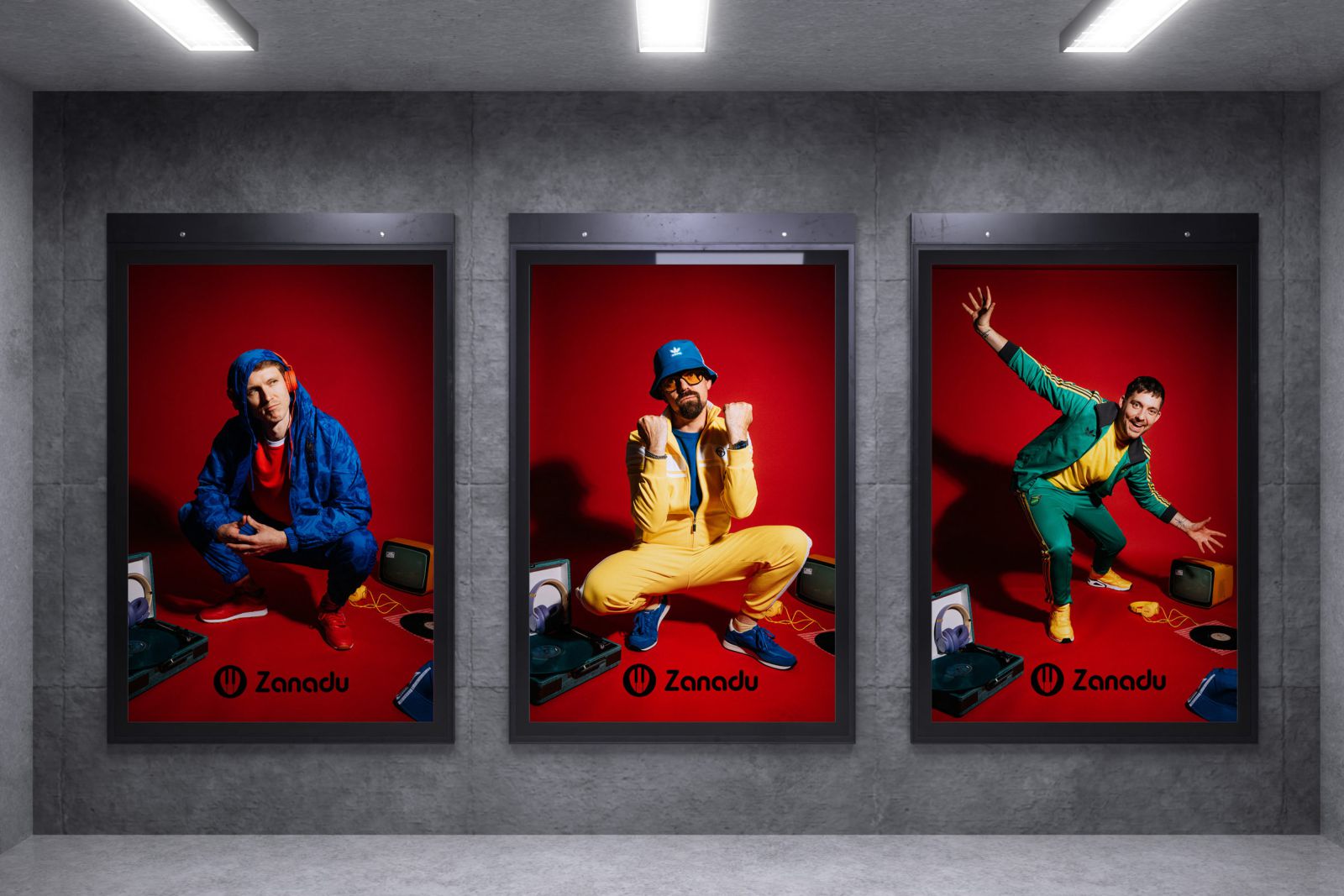 Drei Plakate an einer grauen Wand zeigen Männer in farbenfrohen Outfits - blau, gelb und grün -, die energisch vor roten Hintergründen mit Retro-Elektronik posieren. Jedes Poster zeigt das Zanadu-Logo am unteren Rand.