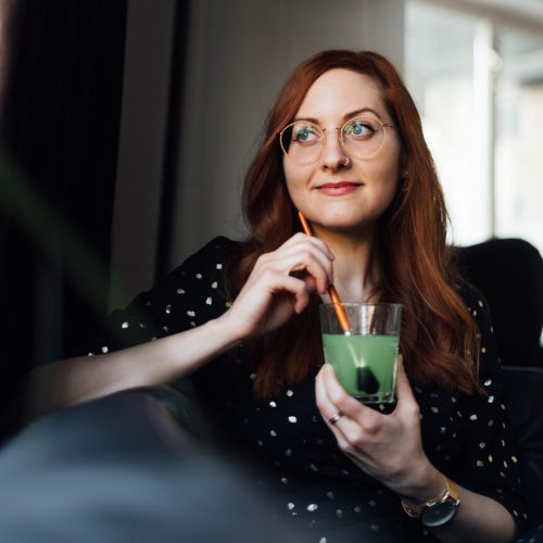 Eine Frau mit langen roten Haaren und Brille, die ein schwarzes Kleid mit weißen Punkten trägt, sitzt in einem Haus und hält ein Glas mit einem grünen Getränk mit einem Strohhalm in der Hand und schaut nachdenklich zur Seite. Im Hintergrund ist eine Stehlampe zu sehen.