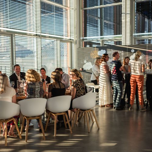 Eine Gruppe von Menschen sitzt an einem Tisch und diskutiert, während andere in einem hellen, modernen Raum mit großen Fenstern und Sonnenlicht, das durch die Jalousien fällt, daneben stehen und sich unterhalten.