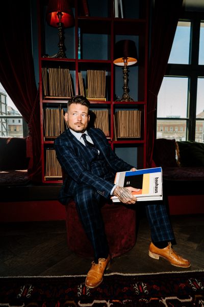 Ein Mann in einem dunkel karierten Anzug sitzt auf einem roten Hocker in einer stilvollen Bibliothek und hält ein Buch in der Hand. Er hat Tätowierungen an der Hand, trägt hellbraune Schuhe und ist von Regalen mit Schallplatten und zwei roten Lampen umgeben. Das Sonnenlicht fällt durch das Fenster.