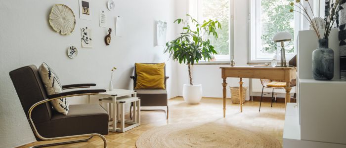 Helles, minimalistisches Wohnzimmer mit Holzfußboden, braunem Sessel, kleinen weißen Tischen, Schreibtisch an großen Fenstern, Topfpflanze, Wandkunst und dekorativen Vasen auf einem Regal. Natürliches Licht füllt den Raum.