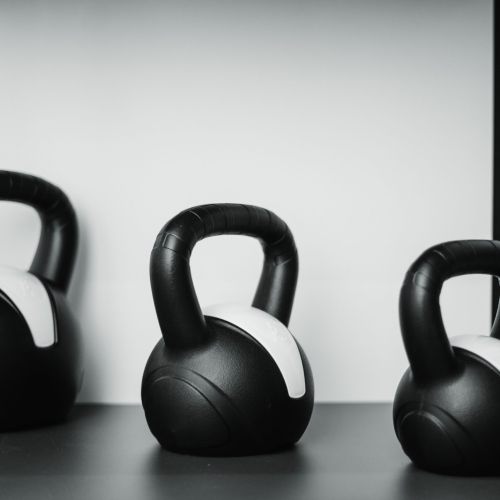 Drei schwarze Kettlebells mit weißen Markierungen befinden sich auf einem dunklen Regal vor einem hellen Hintergrund. Die Kettlebells sind in einer Reihe angeordnet und variieren in ihrer Größe von links nach rechts.