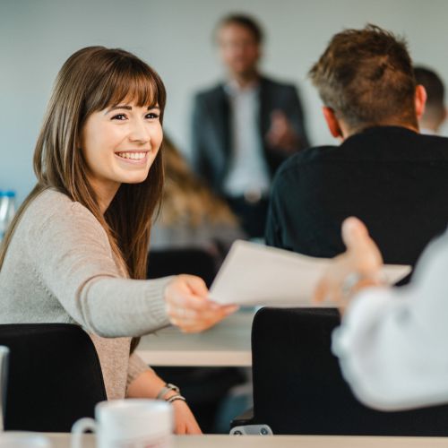 Eine junge Frau, die lächelt und ein Blatt Papier an eine andere Person in einem Klassenzimmer oder einer Besprechung weitergibt, wobei die Teilnehmer sitzen und ein Redner im Hintergrund steht.