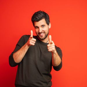 Ein lächelnder Mann mit dunklem Haar und Bart steht vor einem leuchtend roten Hintergrund und zeigt mit beiden Zeigefingern spielerisch in die Kamera. Er trägt ein dunkles langärmeliges Hemd.