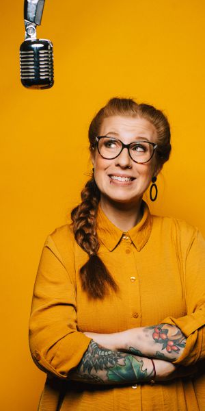 Eine lächelnde Frau mit Brille und geflochtener Frisur steht vor einem gelben Hintergrund und trägt ein gelbes Hemd. Sie ist an den Armen tätowiert und blickt auf ein über ihr hängendes Vintage-Mikrofon.